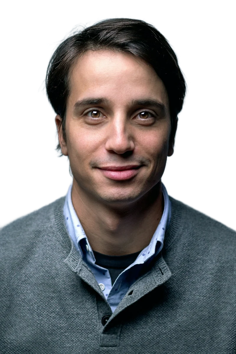 A smiling man in a gray jacket against a muted blue background, background removed